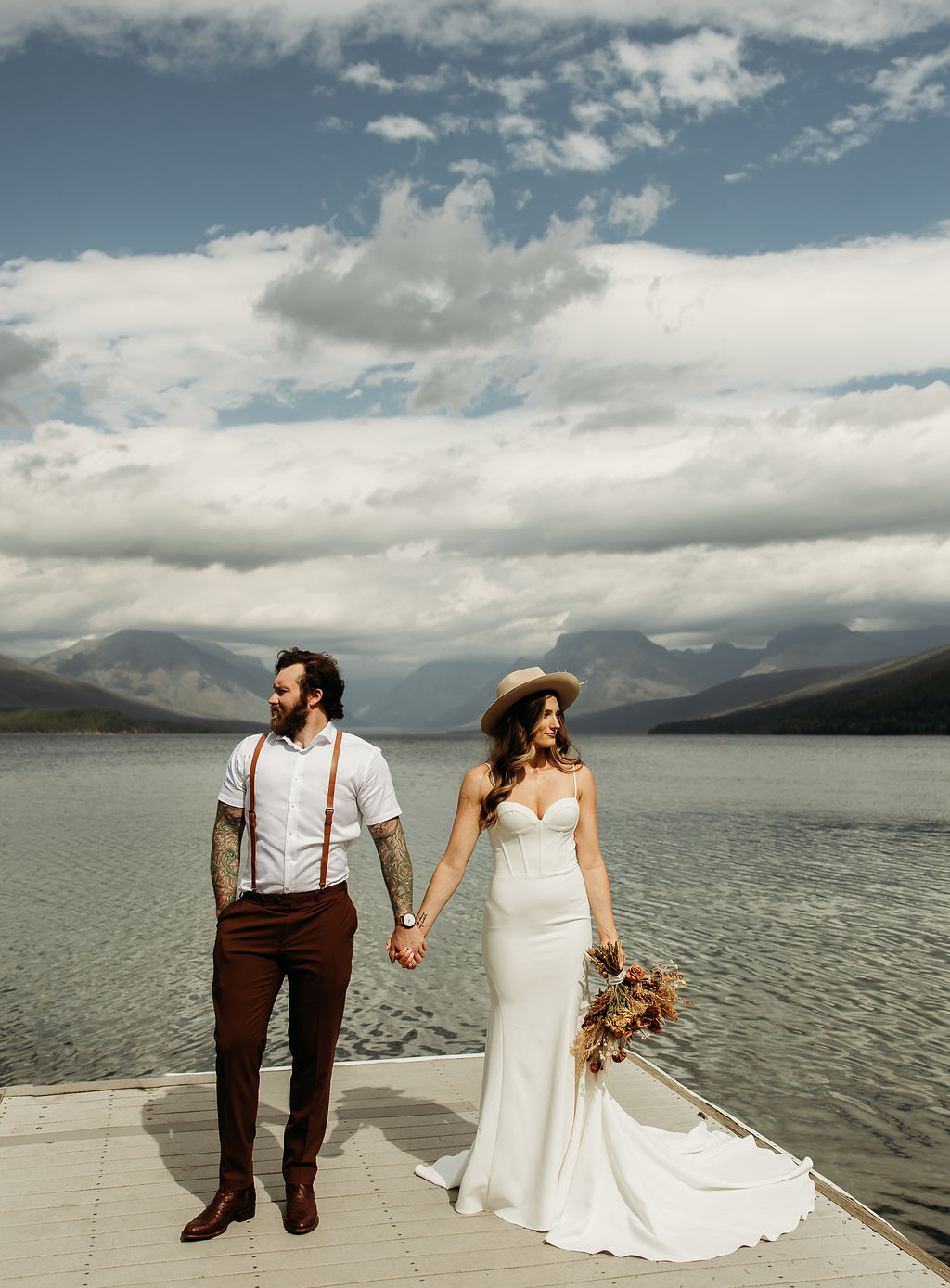 A couple stands on a dock by a lake; the man, in a white shirt and sunglasses, gently touches the woman's face. She holds a bouquet and wears a white dress and hat. Mountains and water are in the background.