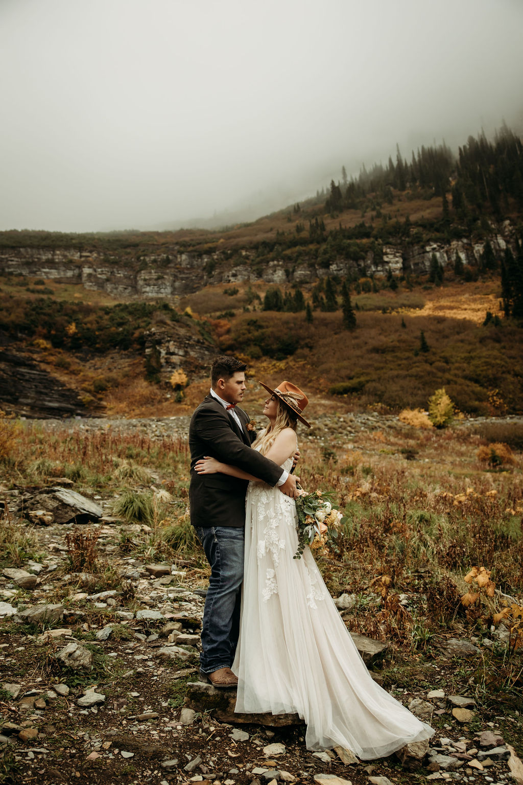 A couple, dressed in wedding attire, embrace and kiss on a rocky outcrop with a mountainous landscape in the background. The sky is overcast and the scene is surrounded by autumnal trees.