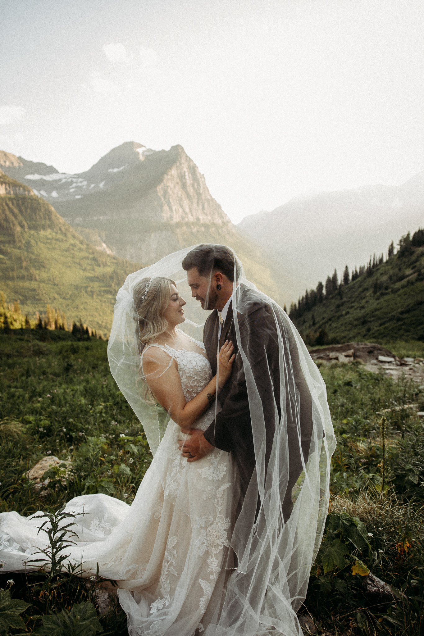 A bride in a white gown and veil holding flowers stands next to a groom in a black suit on a rocky ledge with mountains and valley in the background at their destination elopement