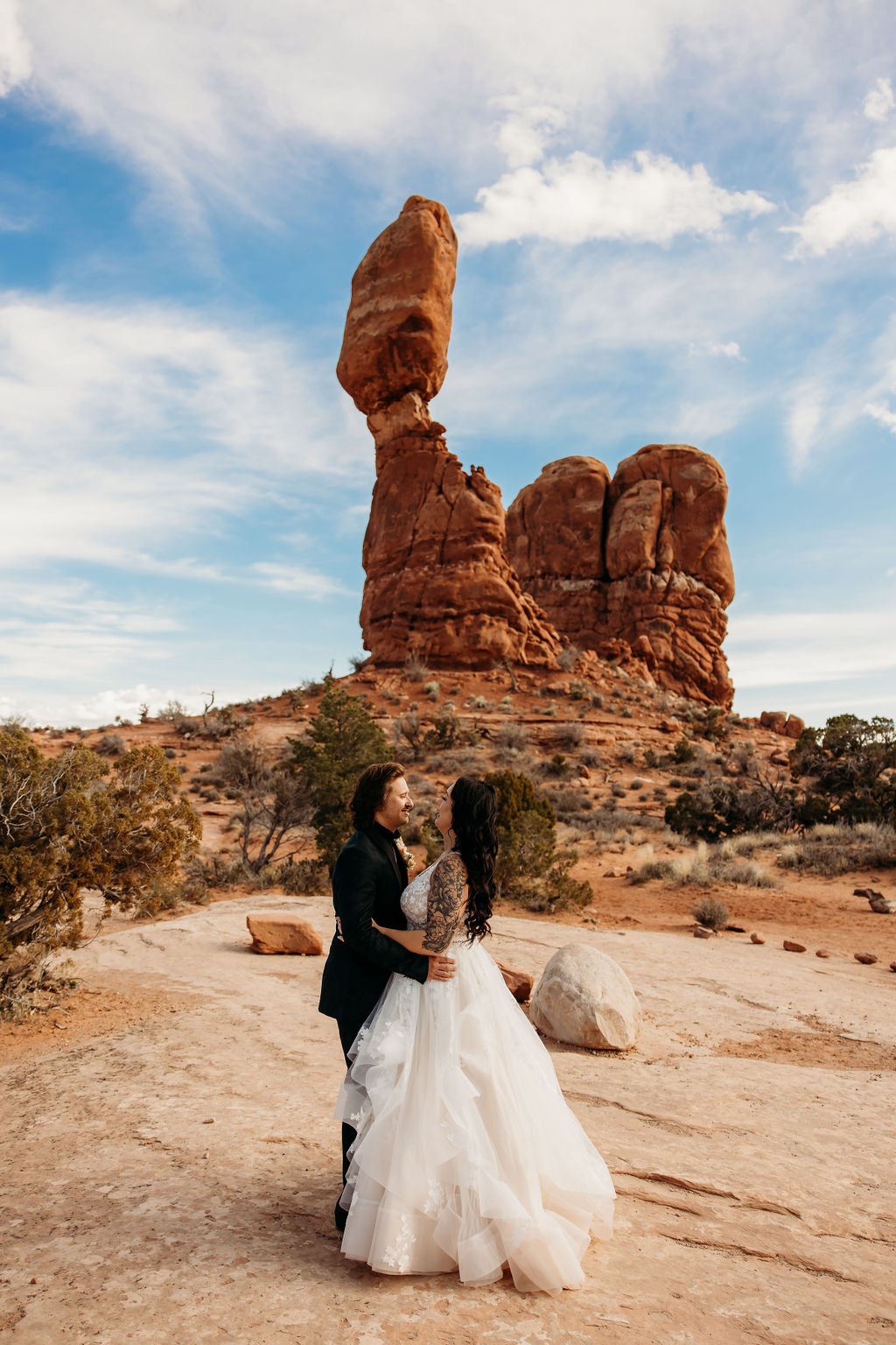 A couple in wedding attire embracing in front of the balanced rock formation in arches national park. | PHOTOGRAPHY BY BROGAN |  5 REASONS TO HAVE A MOAB NATIONAL PARK ELOPEMENT 