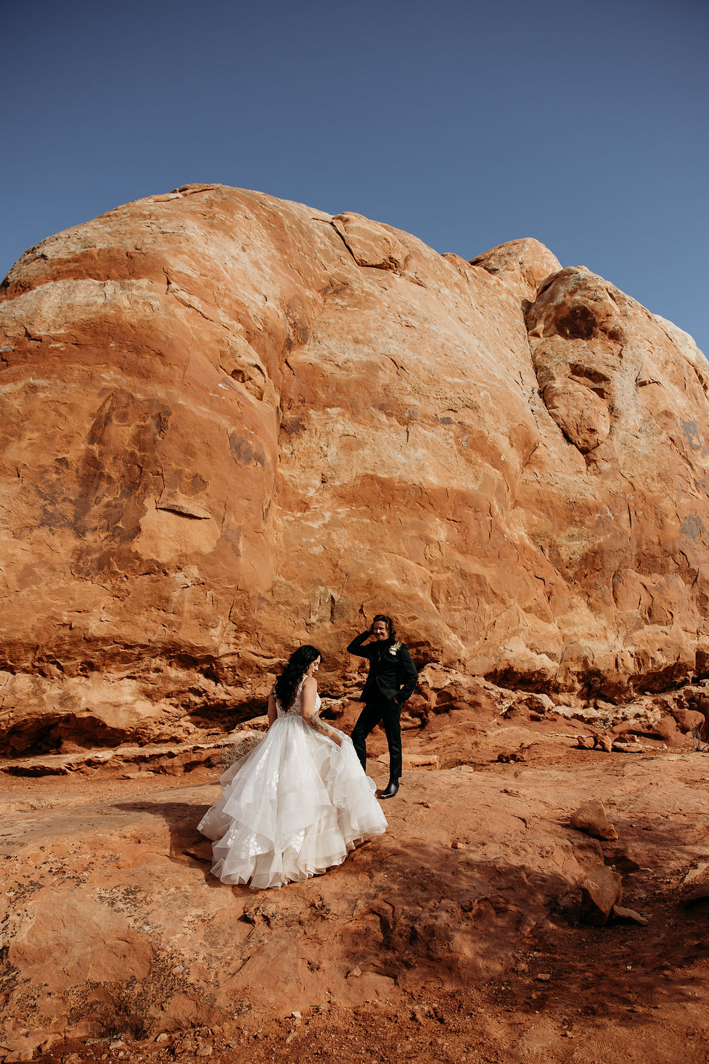 A couple holds hands while walking near a towering rock formation under a clear sky at Arches National Park | PHOTOGRAPHY BY BROGAN |  5 REASONS TO HAVE A MOAB NATIONAL PARK ELOPEMENT 