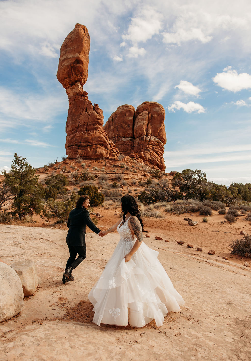 A couple holds hands while walking near a towering rock formation under a clear sky. | PHOTOGRAPHY BY BROGAN |  5 REASONS TO HAVE A MOAB NATIONAL PARK ELOPEMENT 