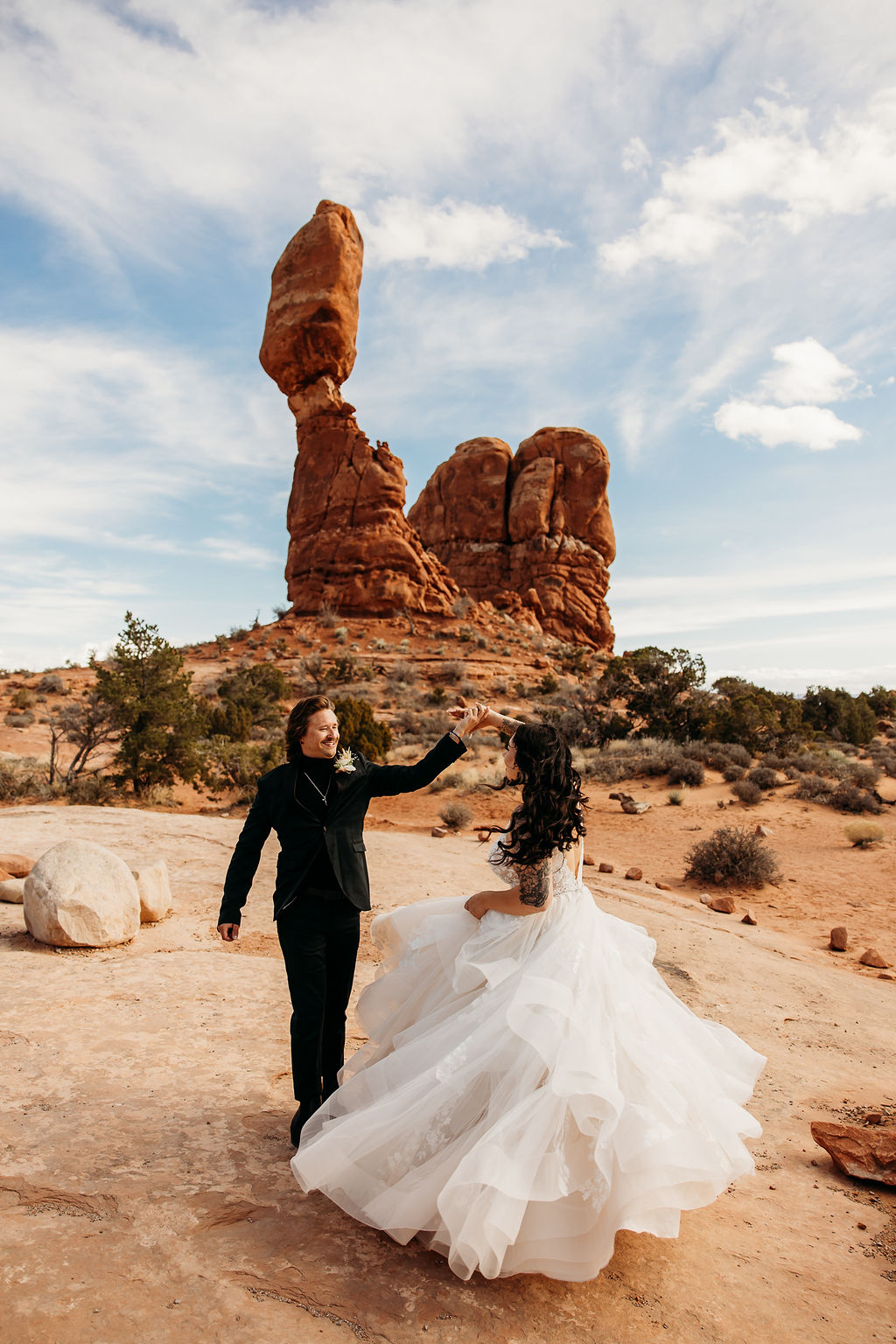 A couple dressed in wedding attire dance in front of a large, unique rock formation in a desert landscape under a partly cloudy sky at their destination elopement