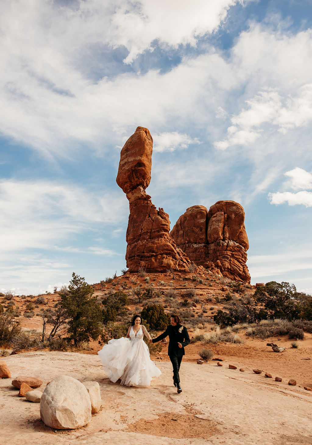 A couple in wedding attire walking hand in hand near a towering rock formation under a cloudy sky. | PHOTOGRAPHY BY BROGAN |  5 REASONS TO HAVE A MOAB NATIONAL PARK ELOPEMENT 