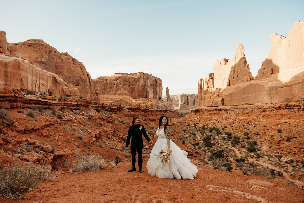 Couple in wedding attire posing and holding hand  in a desert canyon at Arches National park. 
