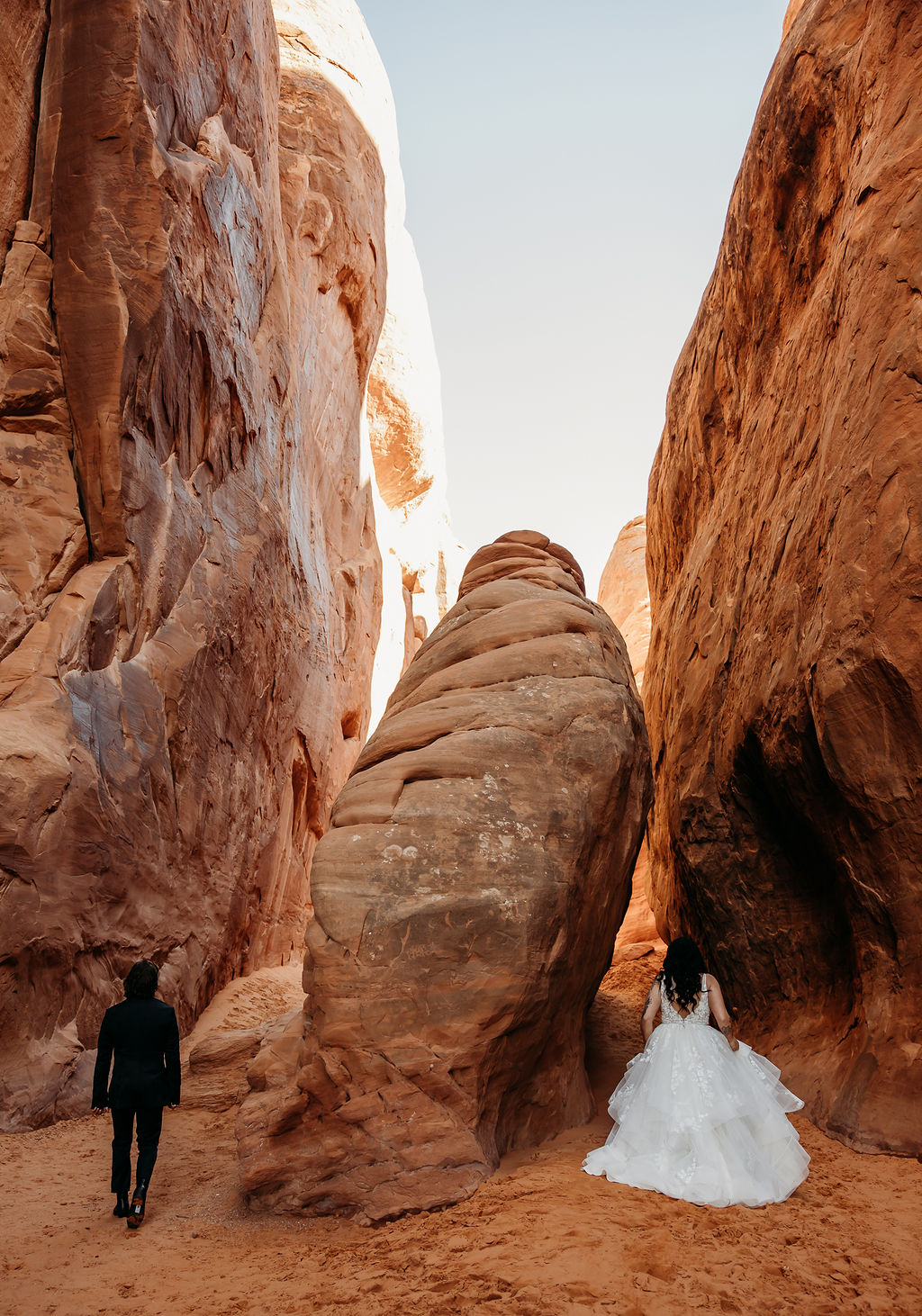A bride and groom walk through a narrow canyon with towering red rock walls at Arches National Park | PHOTOGRAPHY BY BROGAN |  5 REASONS TO HAVE A MOAB NATIONAL PARK ELOPEMENT 