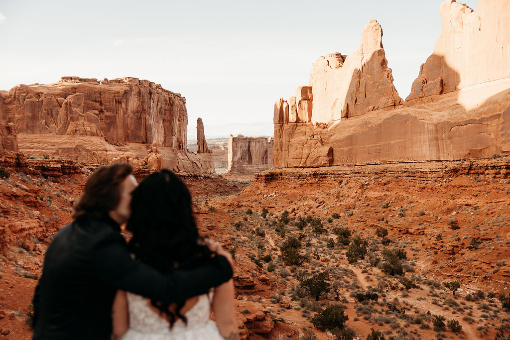 A couple in wedding attire embracing in front of the balanced rock formation. | PHOTOGRAPHY BY BROGAN |  5 REASONS TO HAVE A MOAB NATIONAL PARK ELOPEMENT 