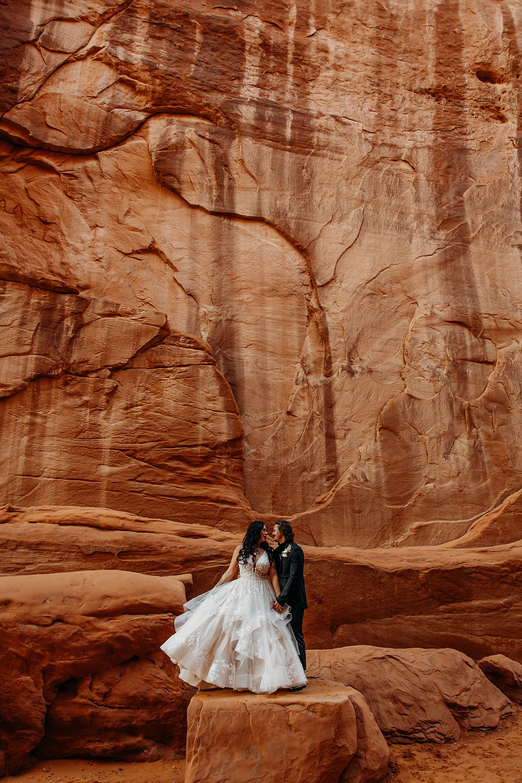A couple holds hands while walking near a towering rock formation under a clear sky. | PHOTOGRAPHY BY BROGAN |  5 REASONS TO HAVE A MOAB NATIONAL PARK ELOPEMENT 