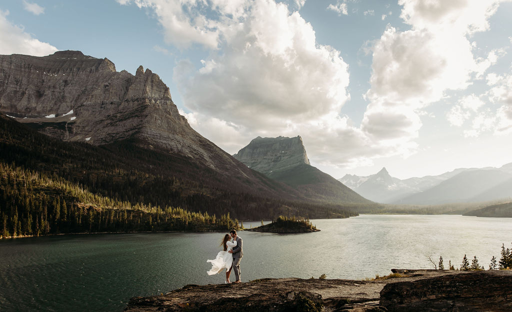 A couple walks hand in hand along a scenic lakeside trail with towering mountains in the background at Glacier National park