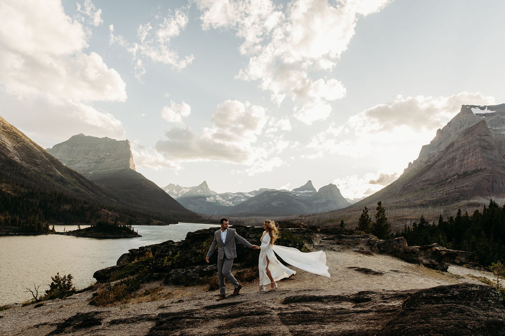 A couple in formal attire walks hand in hand on a rocky landscape with a scenic backdrop of mountains, a lake, and clouds at their destination elopement