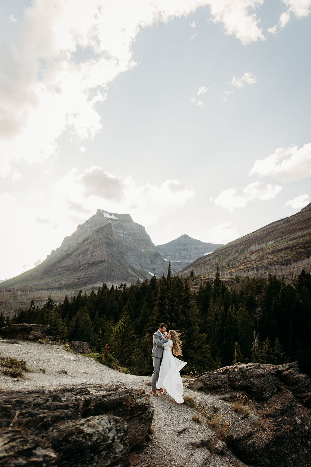 A couple walks hand in hand along a scenic lakeside trail with towering mountains in the background at Glacier National park