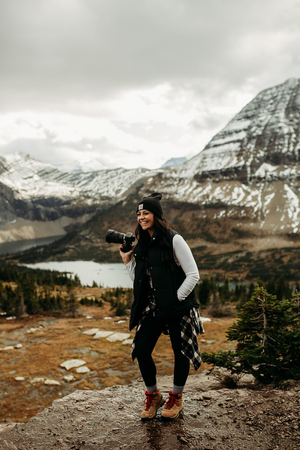 Brogan from photography by brogan in hiking attire smiles while taking photos with a camera in a mountainous landscape with a snowy peak and lake in the background.