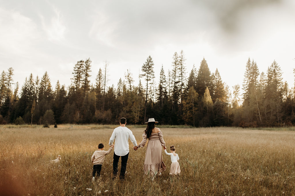 A family of four, including two children, holding hands and walking through a grassy field with a forest in the background.