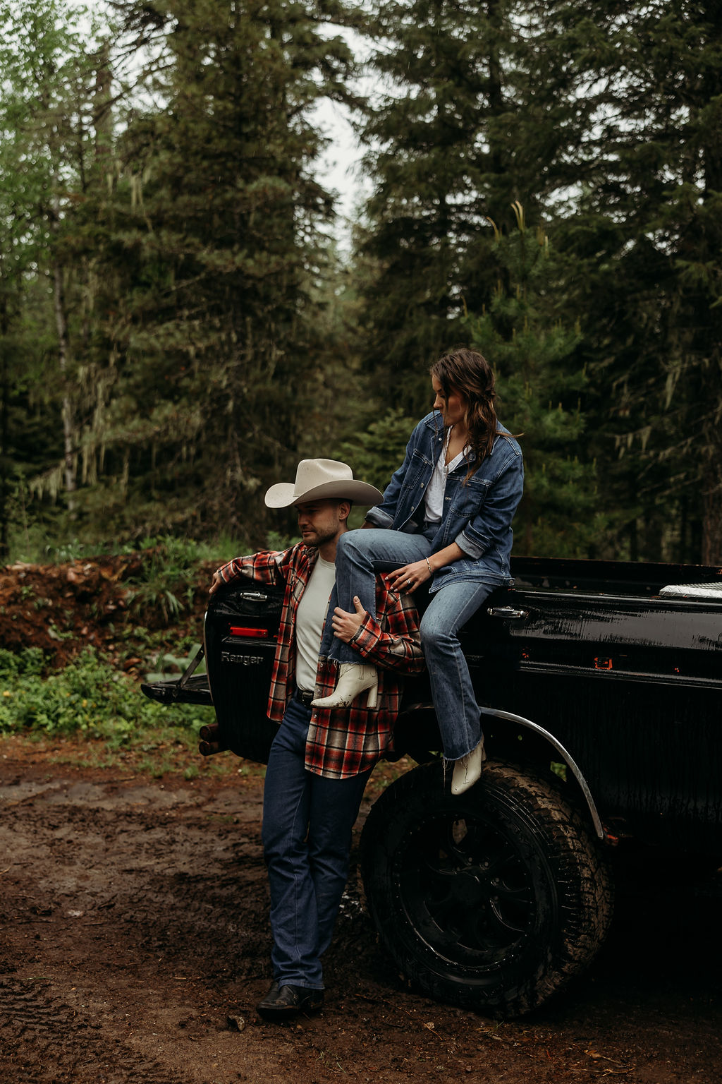 A couple sits close together inside an old, black truck parked in a forested area.