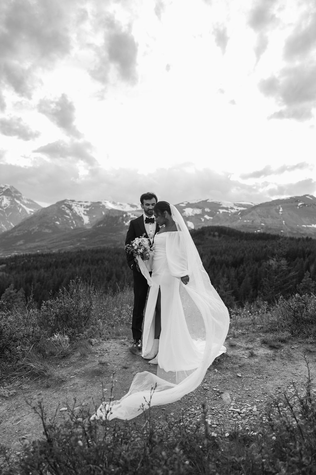 A man and woman stand outside in a grassy area surrounded by trees and mountains. The man hugs the woman from behind, and they both smile. The woman wears a brown dress, and the man wears a cowboy hat during their western engagement photos