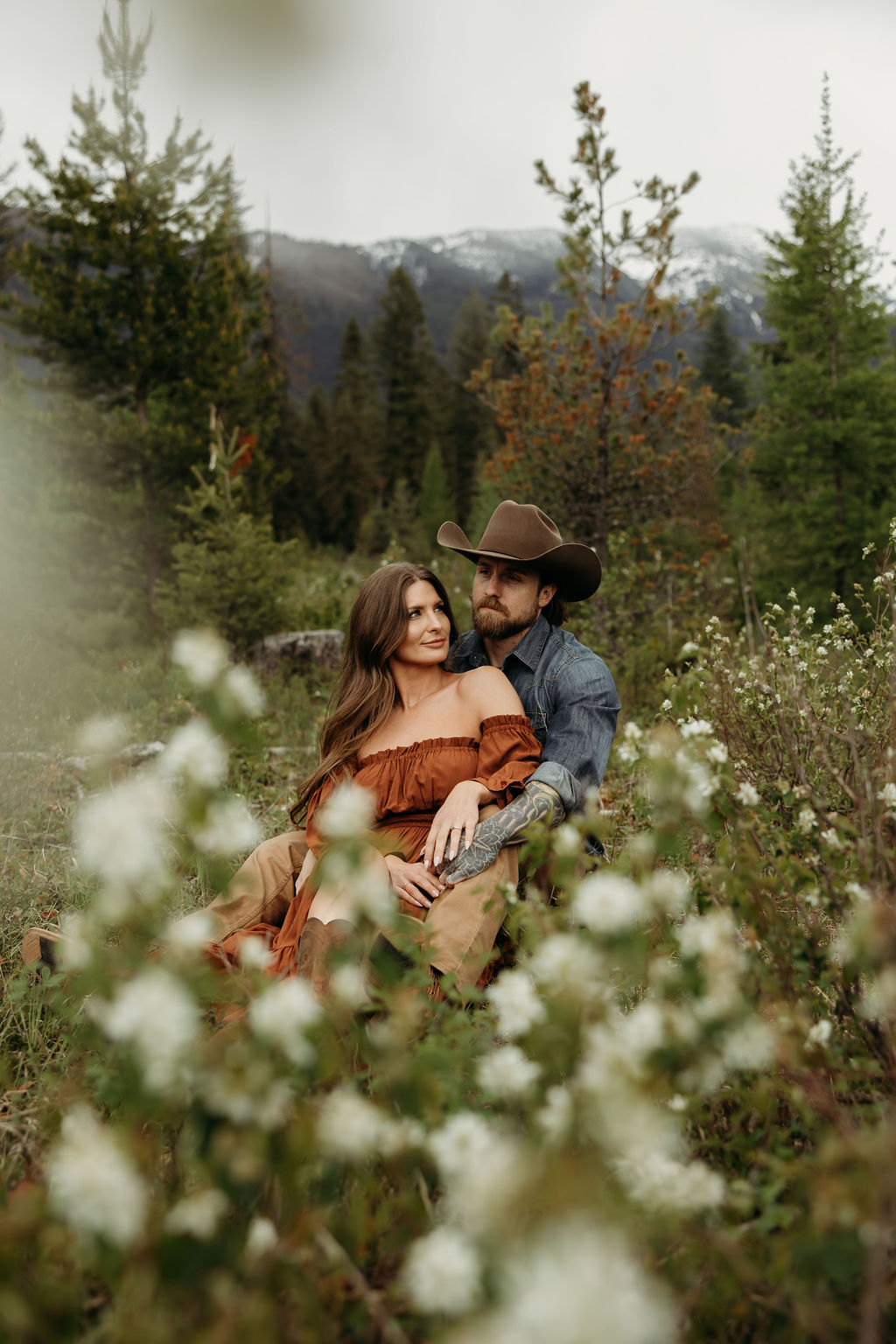 A man and woman stand outside in a grassy area surrounded by trees and mountains. The man hugs the woman from behind, and they both smile. The woman wears a brown dress, and the man wears a cowboy hat during their western engagement photos