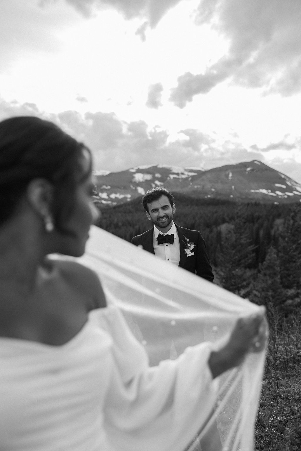 A man and woman stand outside in a grassy area surrounded by trees and mountains. The man hugs the woman from behind, and they both smile. The woman wears a brown dress, and the man wears a cowboy hat during their western engagement photos