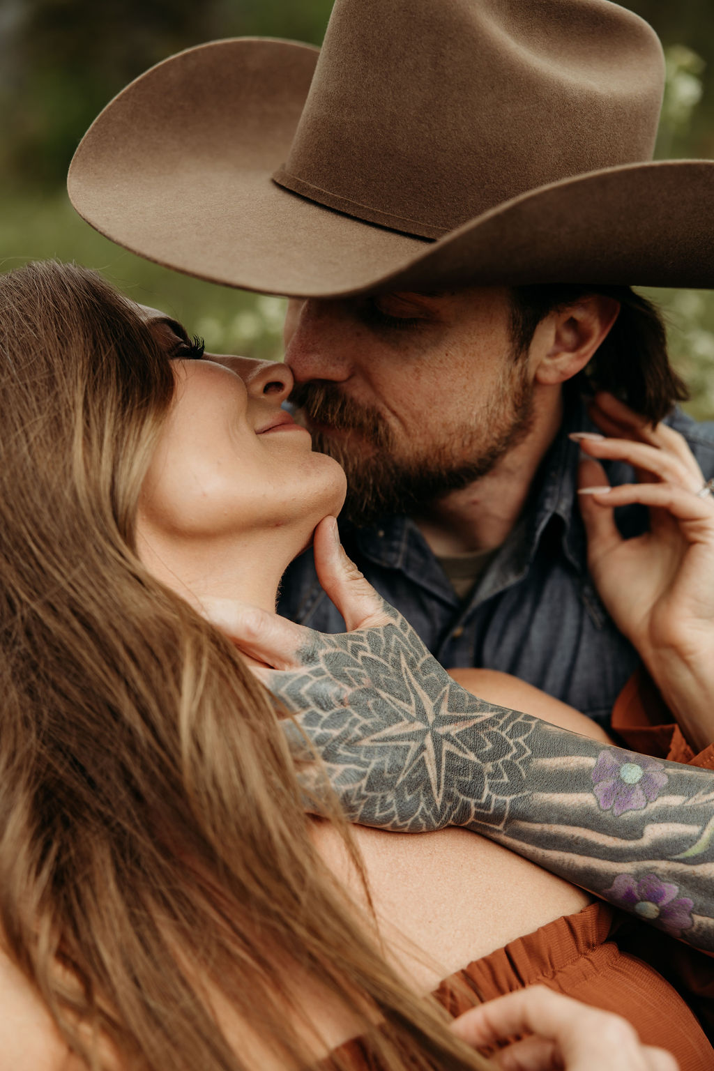 A man and woman stand outside in a grassy area surrounded by trees and mountains. The man hugs the woman from behind, and they both smile. The woman wears a brown dress, and the man wears a cowboy hat during their western engagement photos