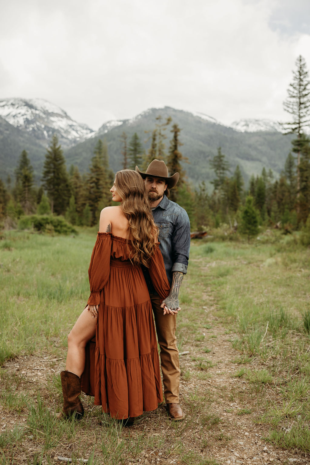 A man and woman stand outside in a grassy area surrounded by trees and mountains. The man hugs the woman from behind, and they both smile. The woman wears a brown dress, and the man wears a cowboy hat during their western engagement photos