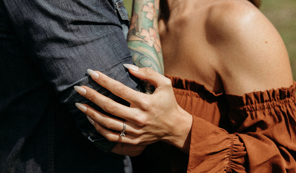 A man and woman stand outside in a grassy area surrounded by trees and mountains. The man hugs the woman from behind, and they both smile. The woman wears a brown dress, and the man wears a cowboy hat during their western engagement photos