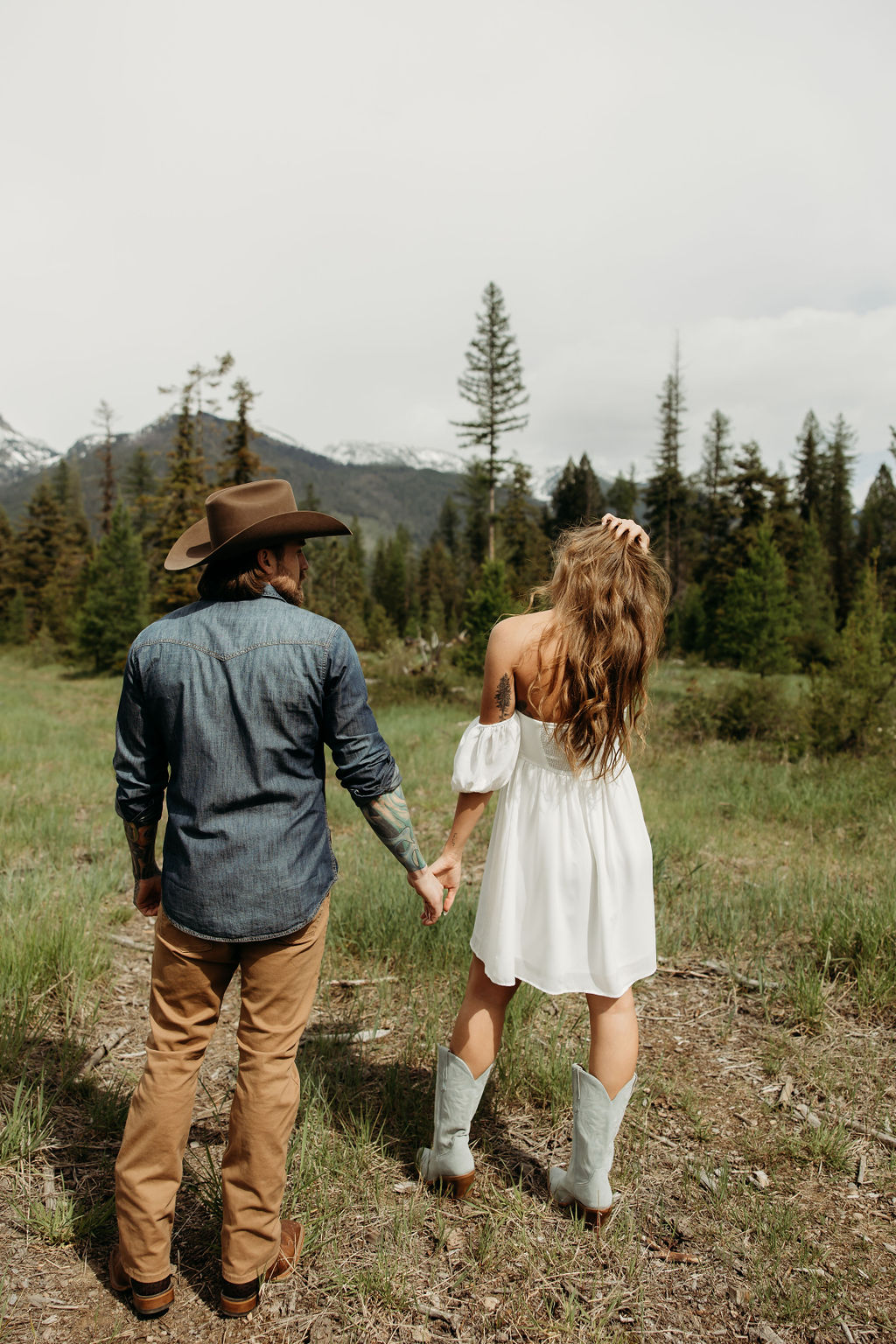 A man in a cowboy hat and jeans playfully holds hands with a woman in a white dress and boots as they walk through a grassy, wooded area during their western engagement photos