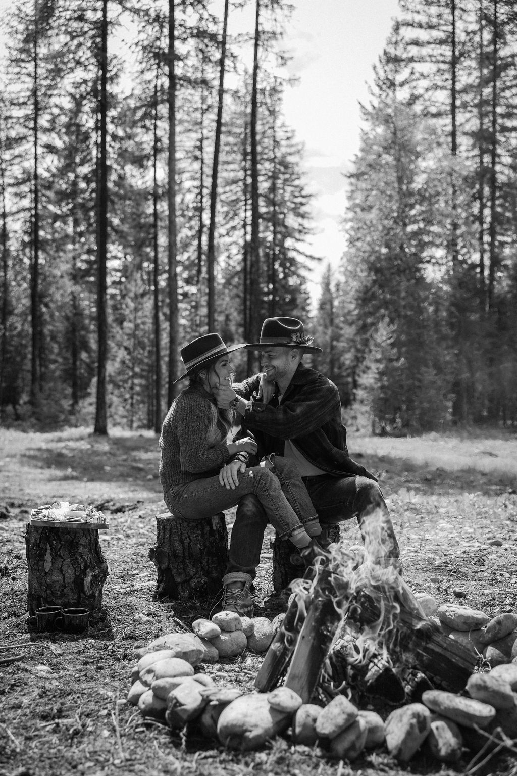 A couple sits on a tree stump in a forest, embracing beside a stone-lined campfire with cooking food. Large trees and green foliage surround the clearing during their western engagement photos