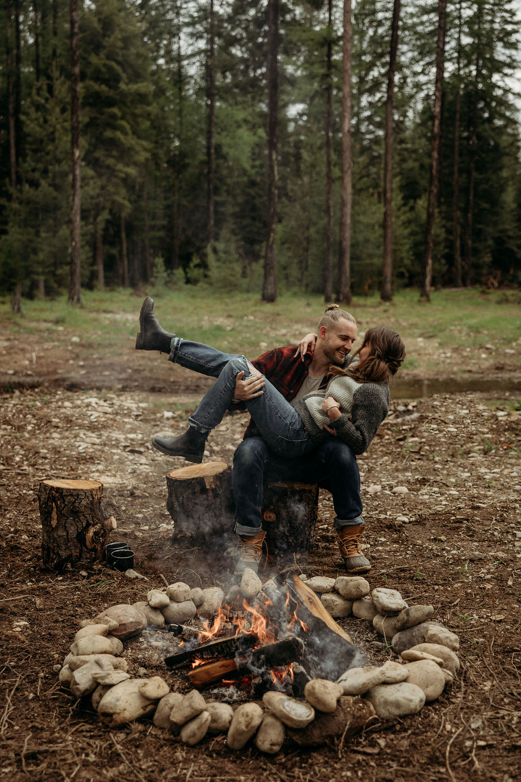 A couple sits on a tree stump in a forest, embracing beside a stone-lined campfire with cooking food. Large trees and green foliage surround the clearing during their western engagement photos