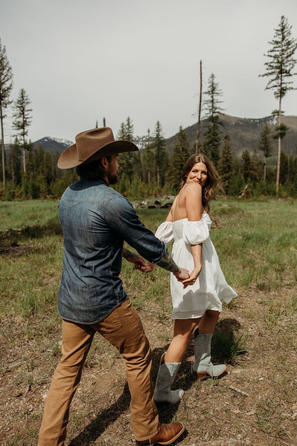 A man in a cowboy hat and jeans playfully holds hands with a woman in a white dress and boots as they walk through a grassy, wooded area during their western engagement photos