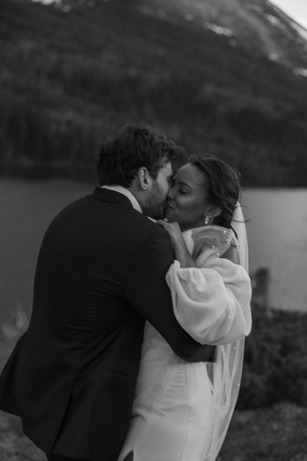 A man in a cowboy hat and jeans playfully holds hands with a woman in a white dress and boots as they walk through a grassy, wooded area during their western engagement photos