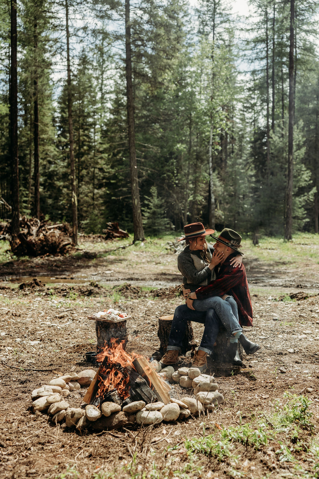 A couple sits on a tree stump in a forest, embracing beside a stone-lined campfire with cooking food. Large trees and green foliage surround the clearing during their western engagement photos