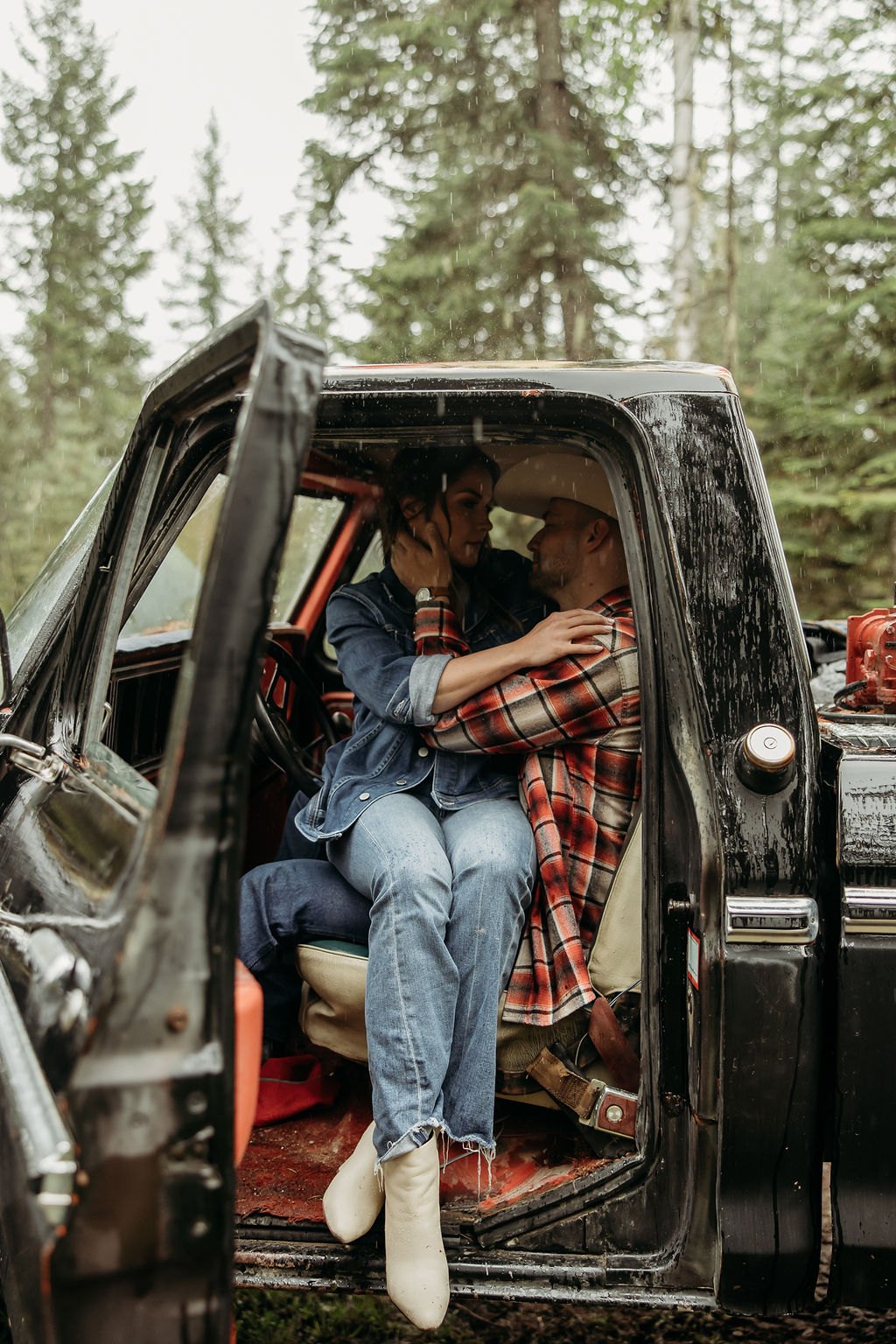 A couple sits close together inside an old, black truck parked in a forested area.