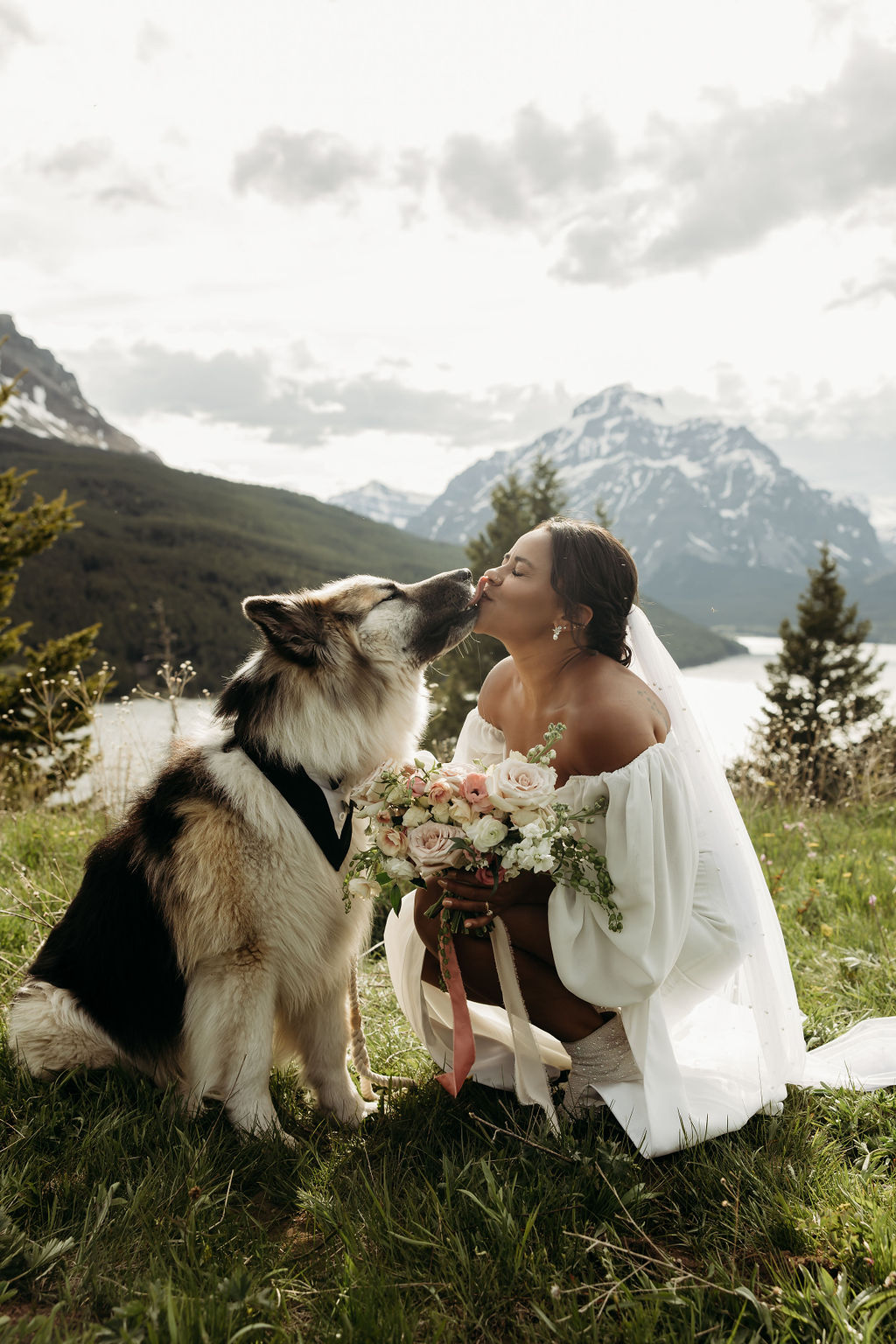 A man and woman stand outside in a grassy area surrounded by trees and mountains. The man hugs the woman from behind, and they both smile. The woman wears a brown dress, and the man wears a cowboy hat during their western engagement photos