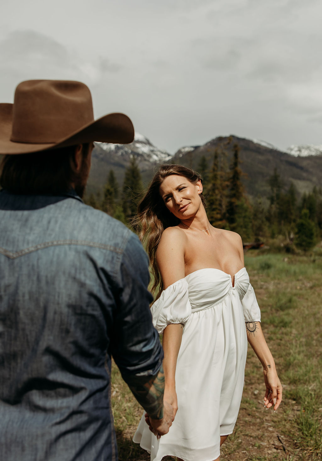 A man in a cowboy hat and jeans playfully holds hands with a woman in a white dress and boots as they walk through a grassy, wooded area during their western engagement photos