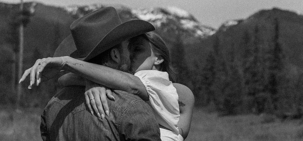 A man in a cowboy hat and jeans playfully holds hands with a woman in a white dress and boots as they walk through a grassy, wooded area during their western engagement photos