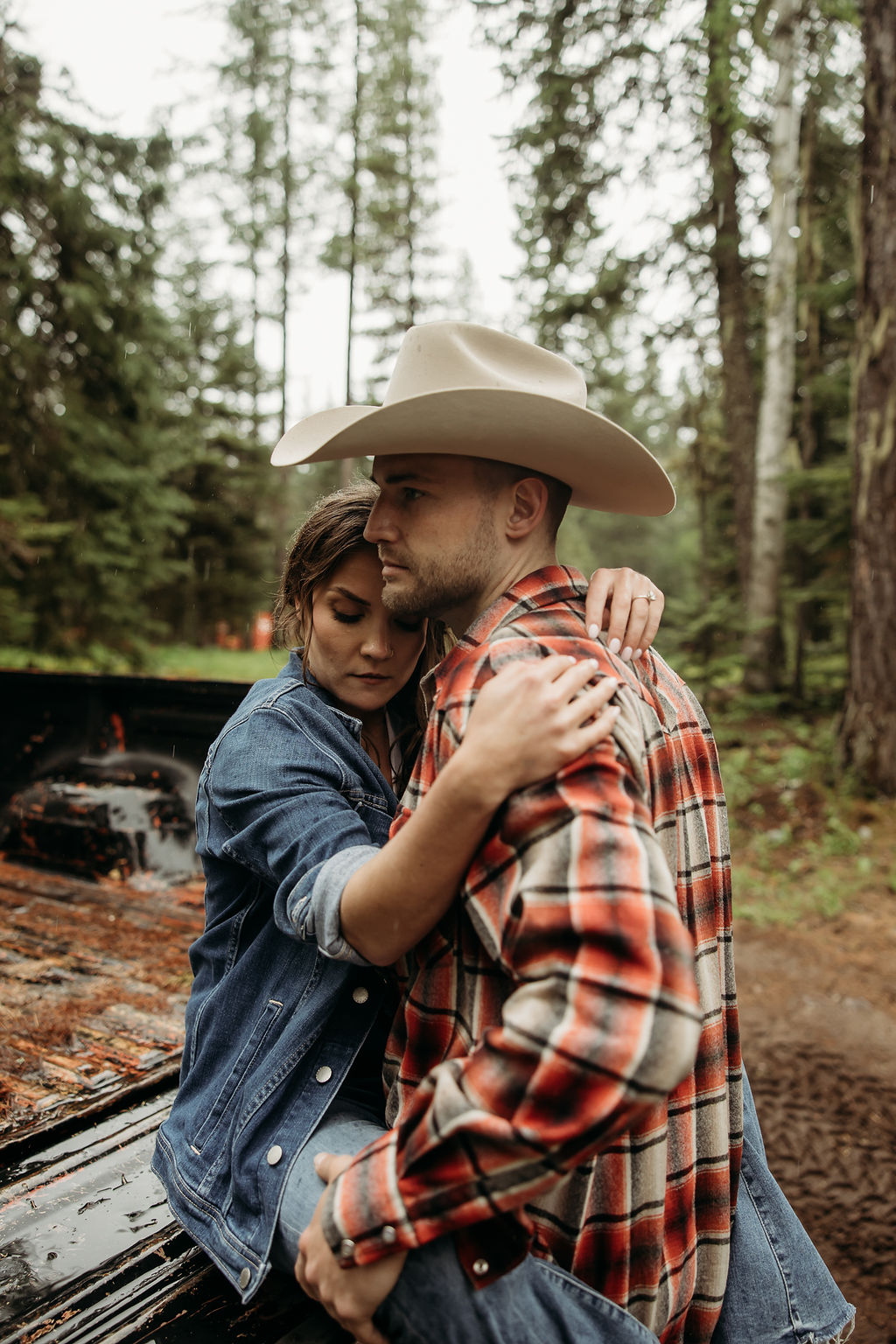 A couple sits close together inside an old, black truck parked in a forested area.