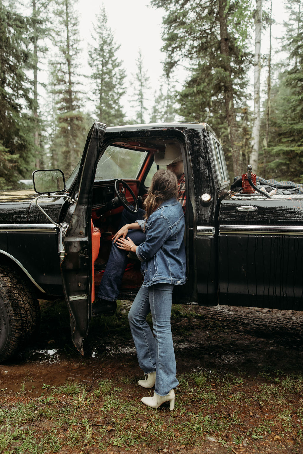 A couple sits close together inside an old, black truck parked in a forested area.