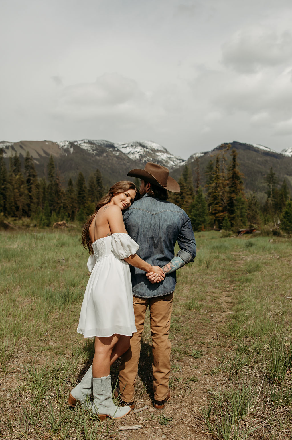 A man in a cowboy hat and jeans playfully holds hands with a woman in a white dress and boots as they walk through a grassy, wooded area during their western engagement photos