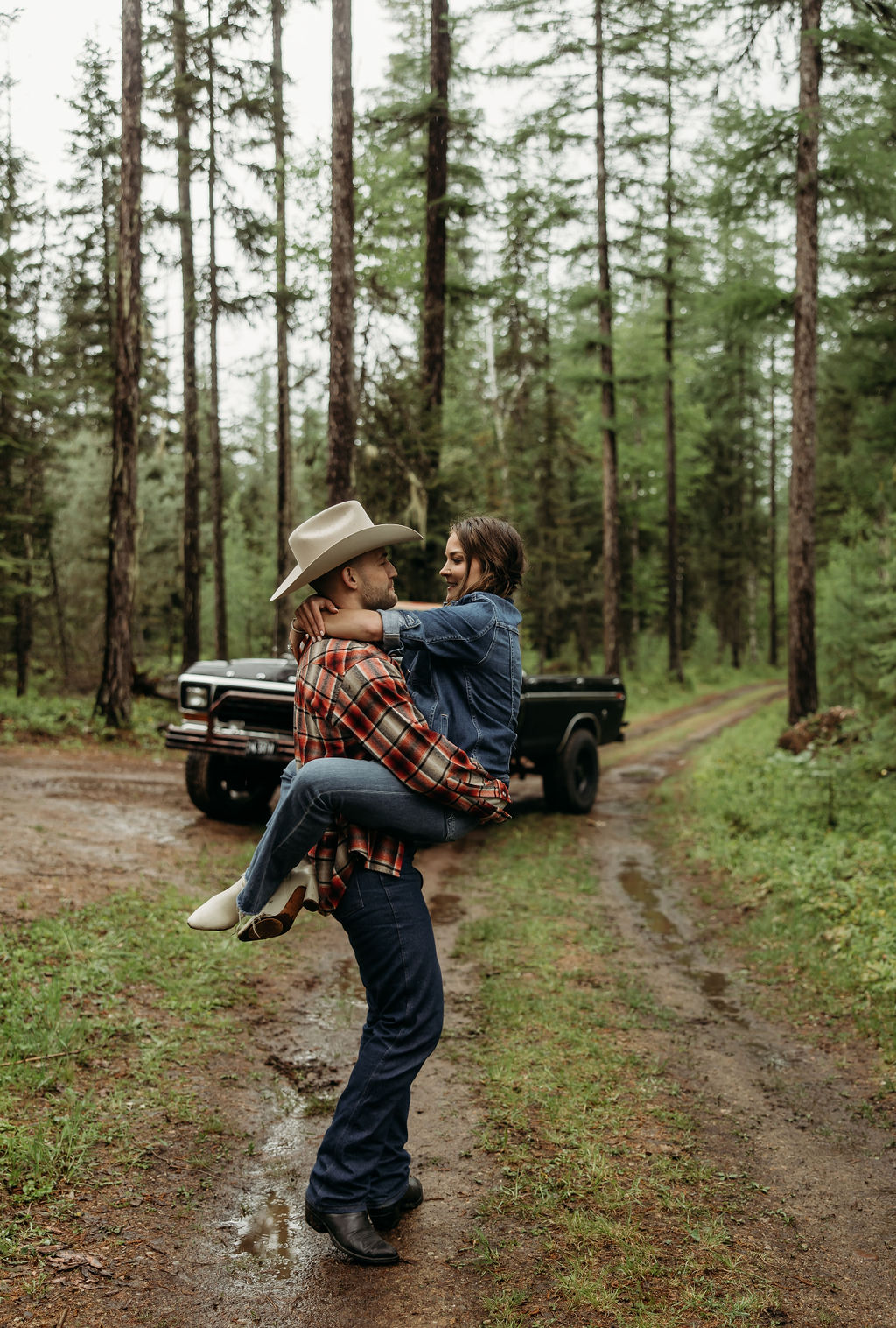 A couple sits close together inside an old, black truck parked in a forested area.