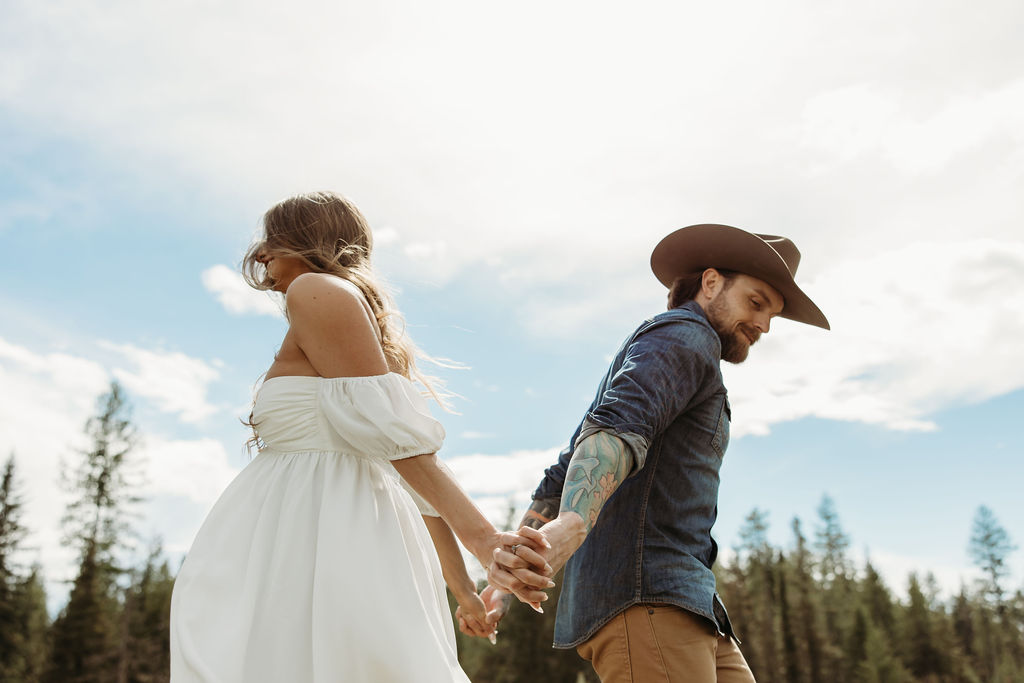 A man in a cowboy hat and jeans playfully holds hands with a woman in a white dress and boots as they walk through a grassy, wooded area during their western engagement photos