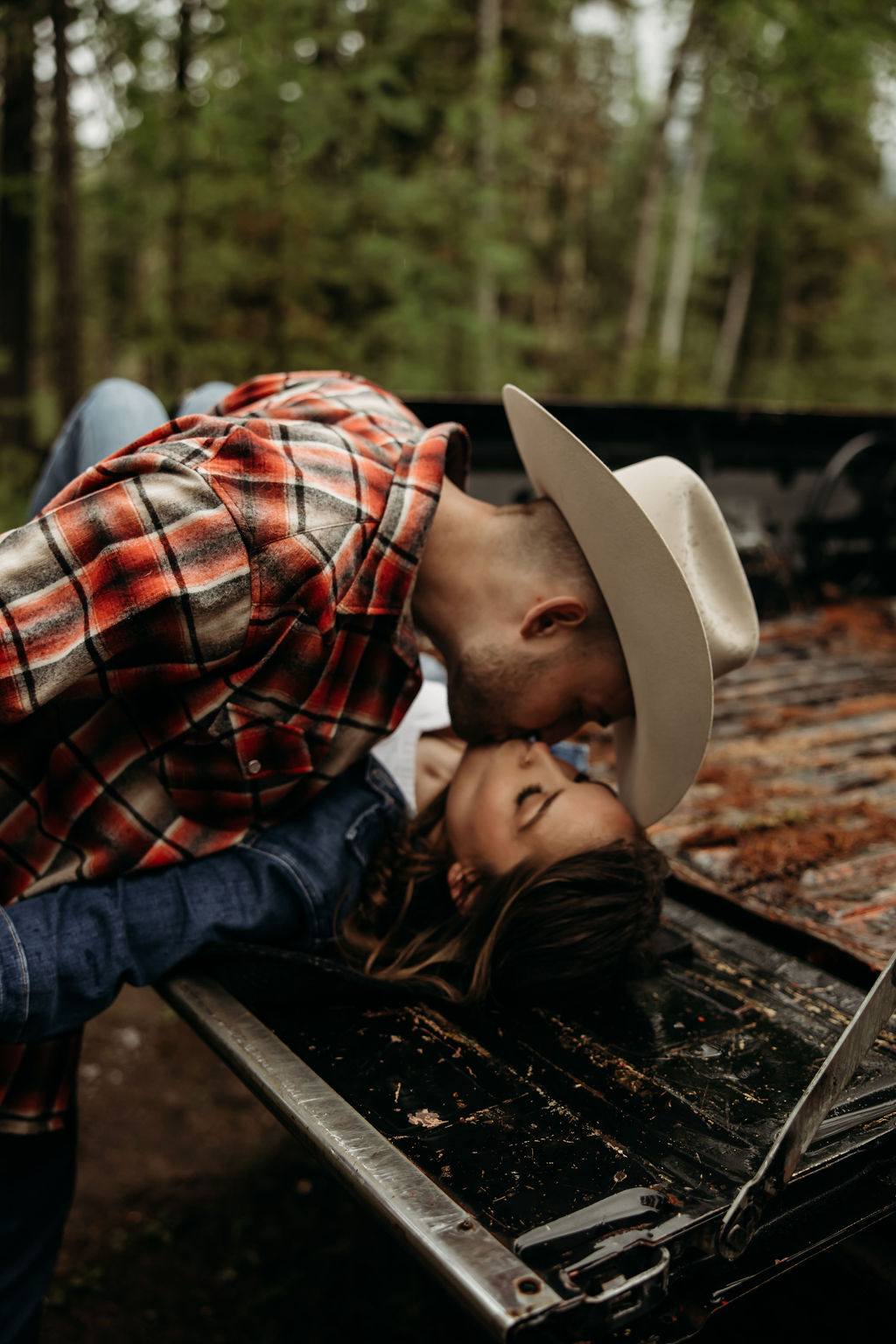 A couple sits close together inside an old, black truck parked in a forested area.