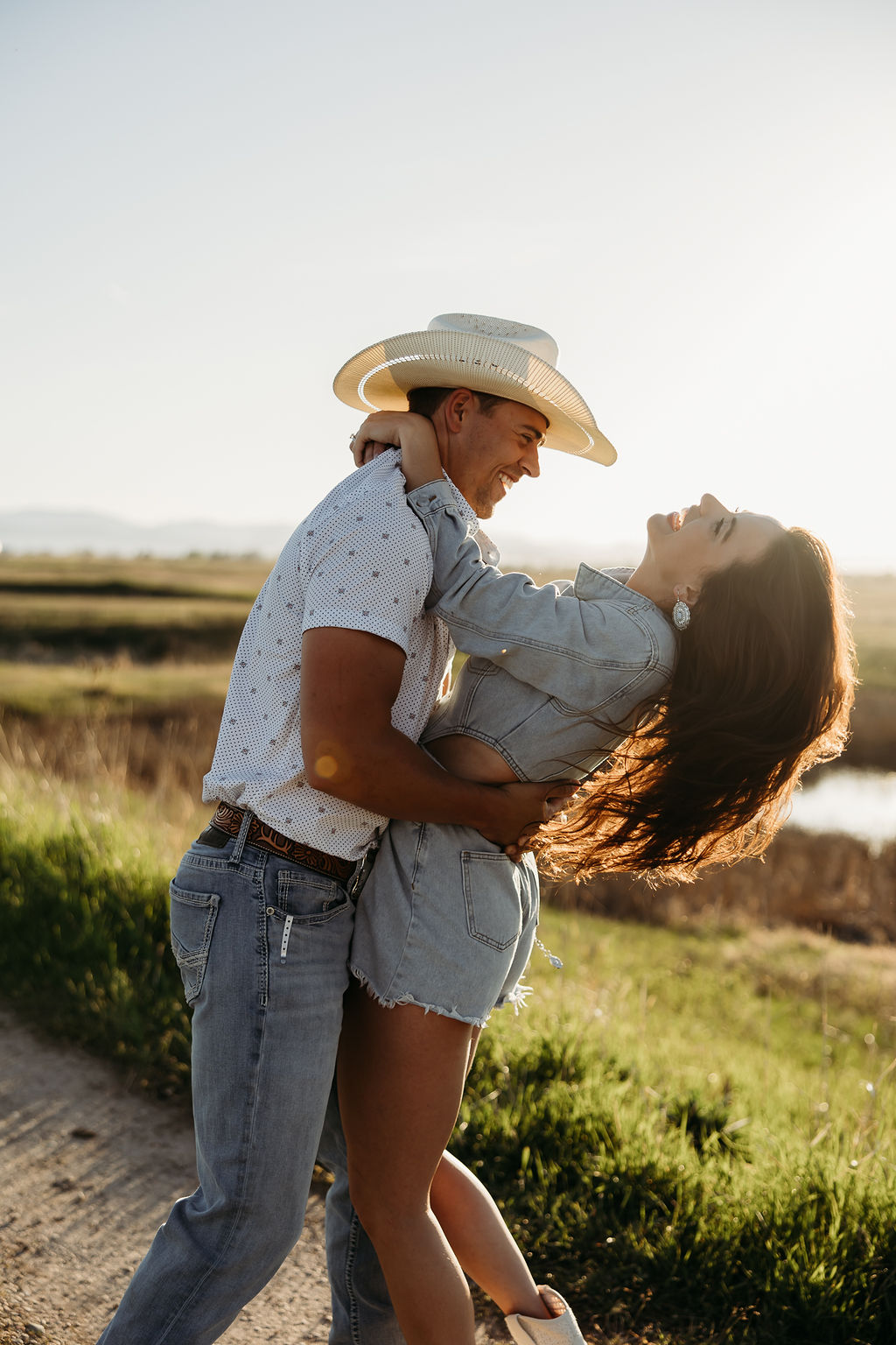 A woman wearing a denim outfit and a man in a cowboy hat stand outdoors with mountains in the background during their western engagement session