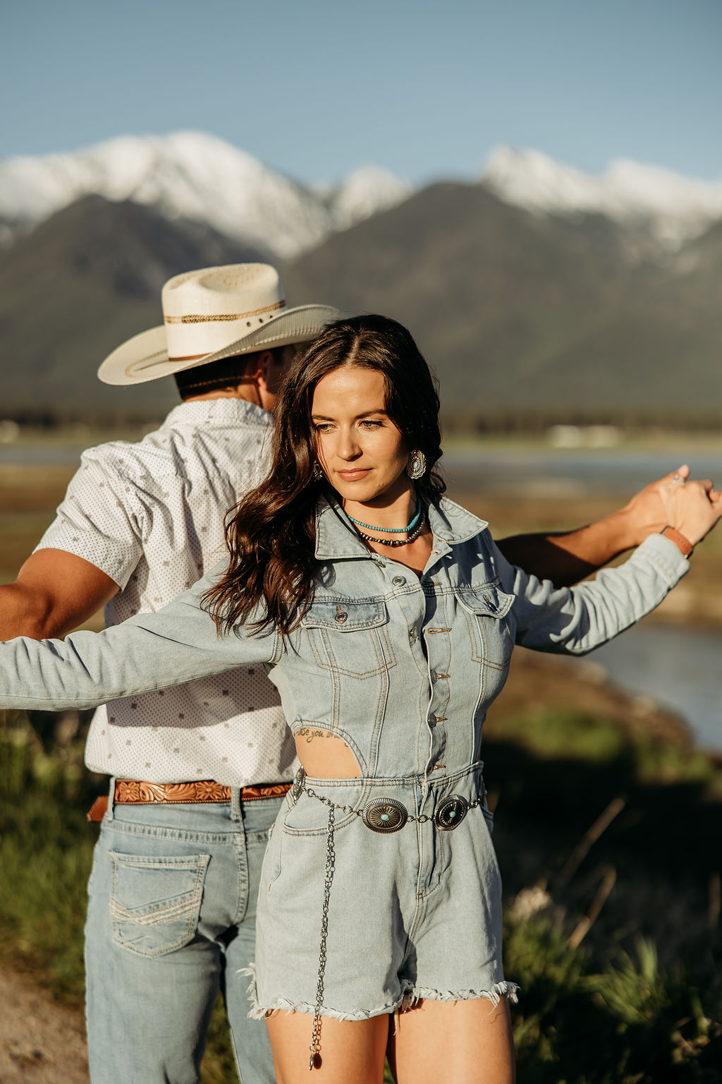 A woman wearing a denim outfit and a man in a cowboy hat stand outdoors with mountains in the background during their western engagement session