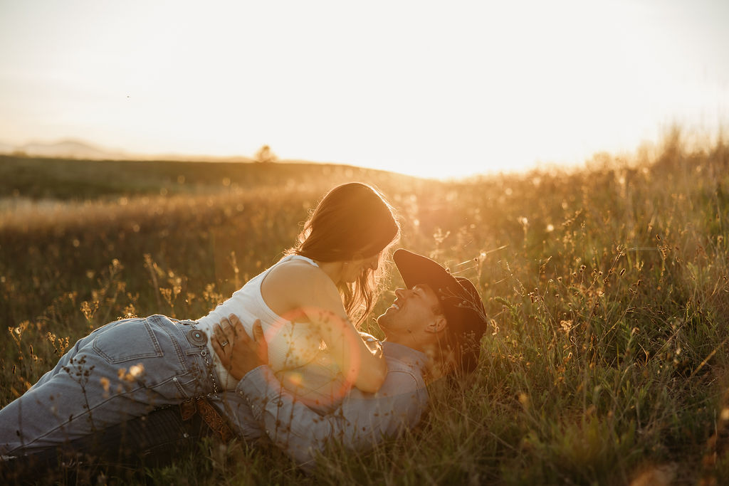 A couple lies in a grassy field at sunset, with one person leaning over the other, both smiling and bathed in warm sunlight.