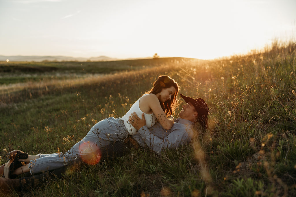 A couple lies in a grassy field at sunset, with one person leaning over the other, both smiling and bathed in warm sunlight.