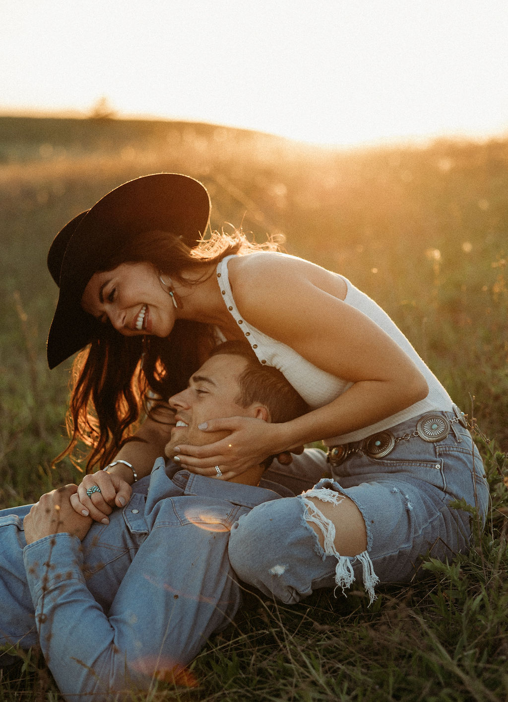 A couple sits in a grassy field at sunset. The woman, laughing and wearing a black hat,. They both wear casual, light-colored clothing during their western engagement photos