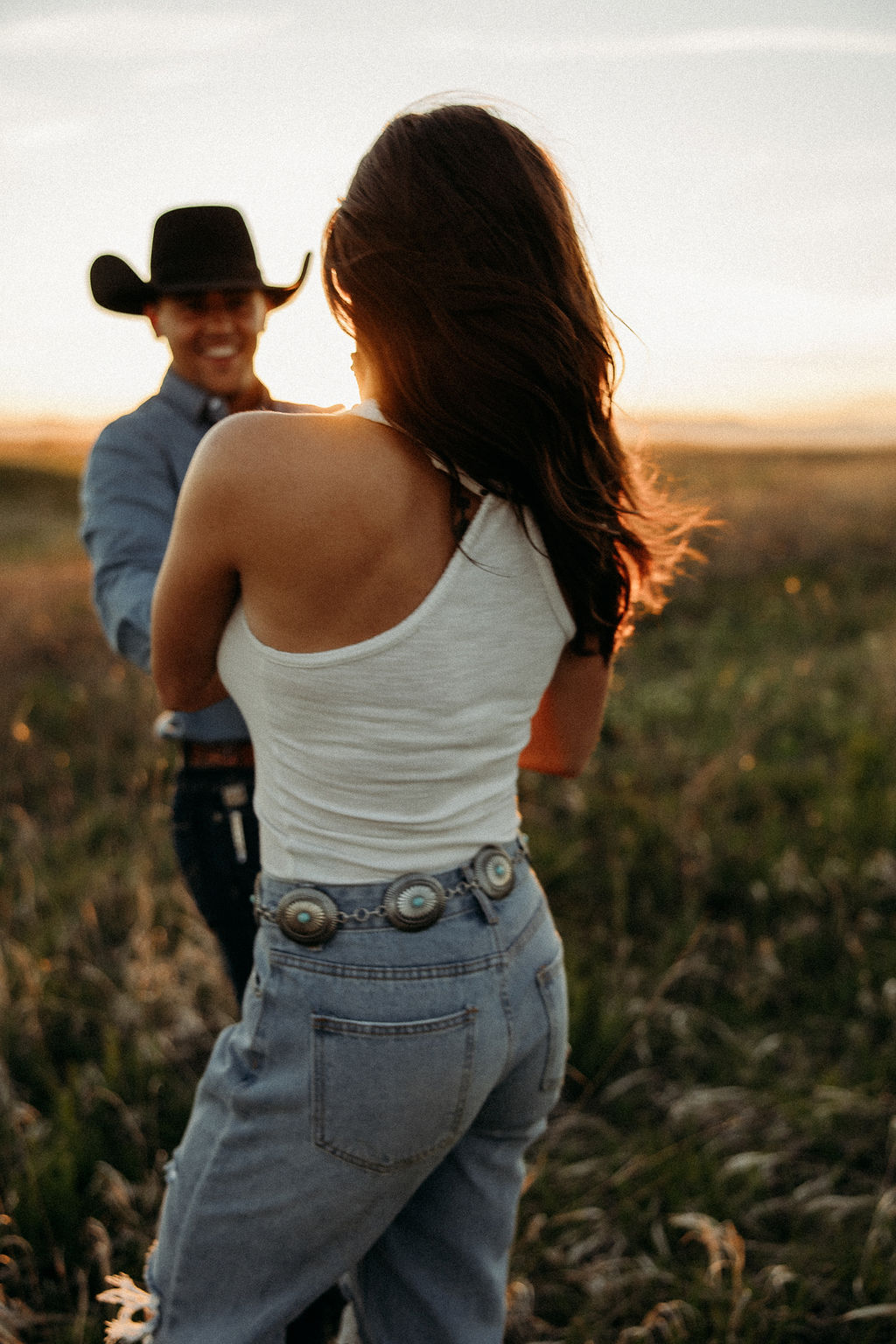 A couple sits in a grassy field at sunset. The woman, laughing and wearing a black hat,. They both wear casual, light-colored clothing during their western engagement photos