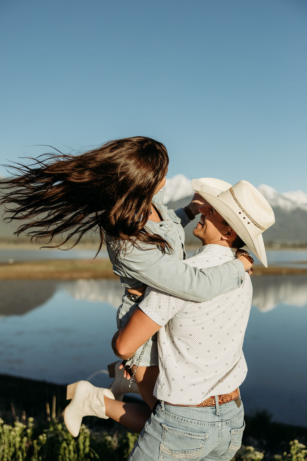 A woman wearing a denim outfit and a man in a cowboy hat stand outdoors with mountains in the background during their western engagement session