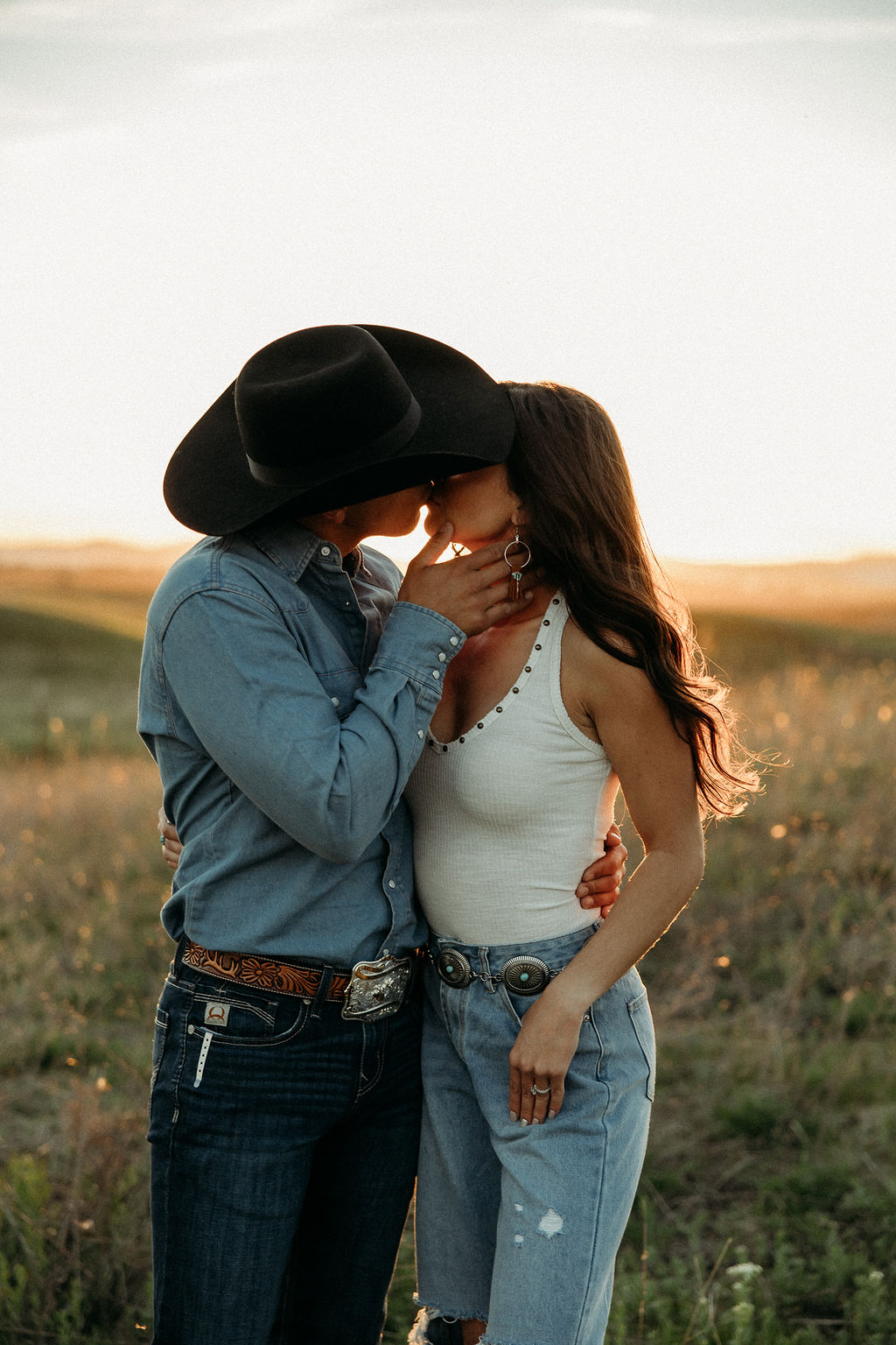 A couple sits in a grassy field at sunset. The woman, laughing and wearing a black hat,. They both wear casual, light-colored clothing during their western engagement photos