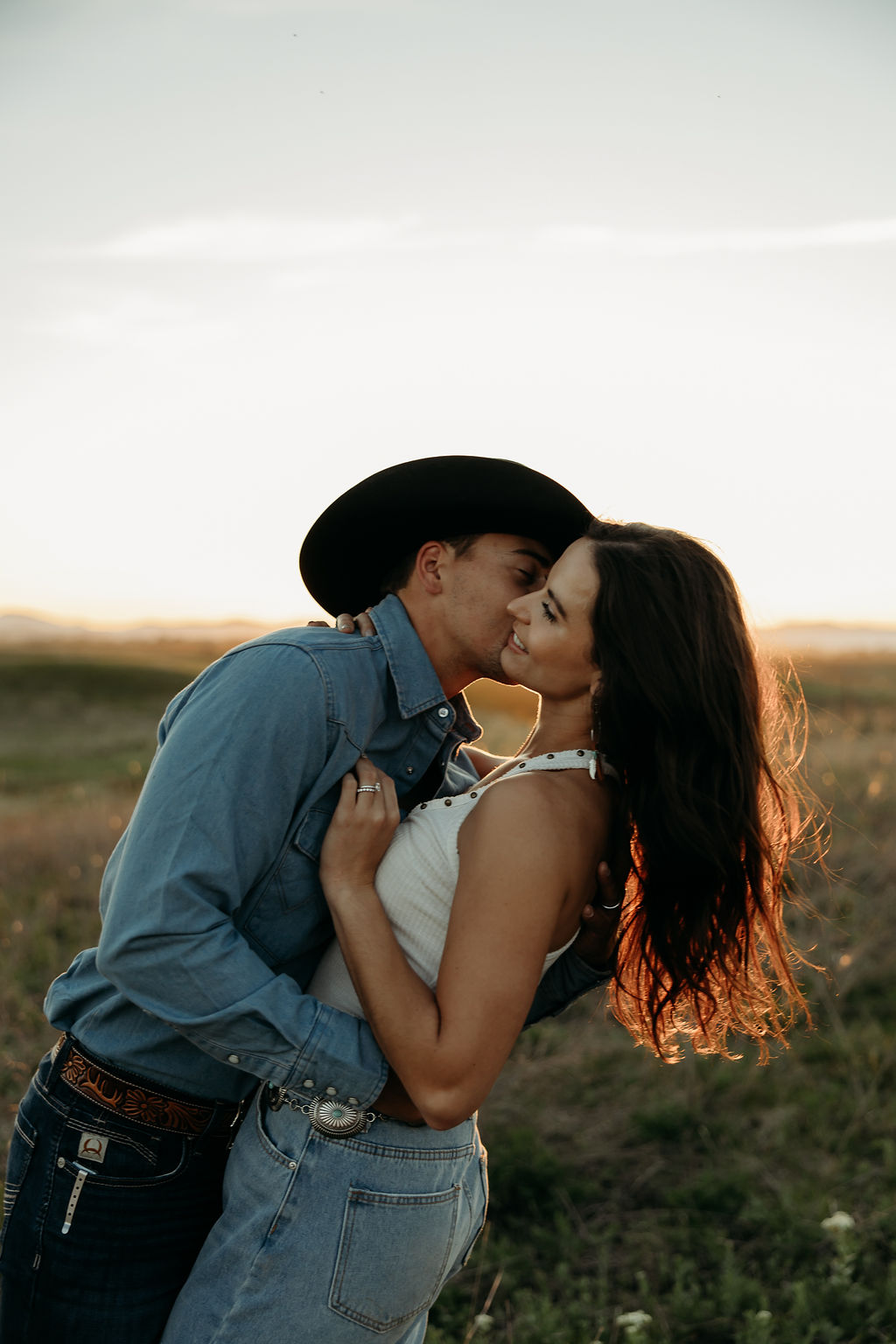 A couple sits in a grassy field at sunset. The woman, laughing and wearing a black hat,. They both wear casual, light-colored clothing during their western engagement photos