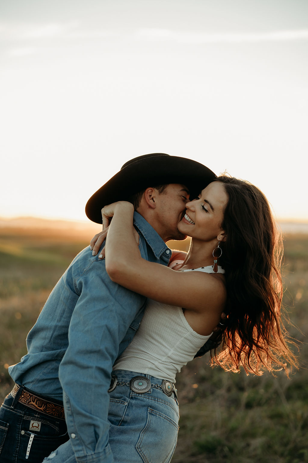 A couple sits in a grassy field at sunset. The woman, laughing and wearing a black hat,. They both wear casual, light-colored clothing during their western engagement photos